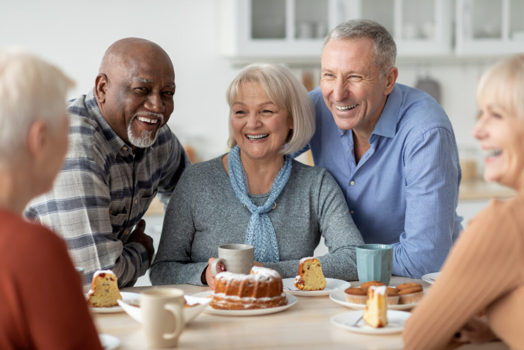 Cheerful senior people of different nationalities sitting at kitchen, drinking tea and eating cake together, having conversation and laughing, chilling together at a senior living community